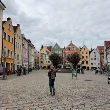Une femme se promène sur la place d'une petite ville européenne pittoresque, bordée de charmants bâtiments colorés. Les arbres dénudés, les boutiques et le ciel nuageux créent une atmosphère paisible et pittoresque.
