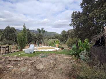 A peaceful garden scene features a covered lounge chair, small pool, lush greenery, trees, mountains in the background, and a cloudy sky on a tranquil day.