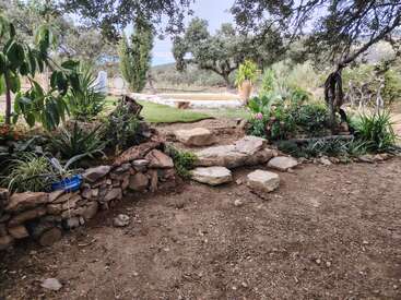 A peaceful garden features stone steps, rustic rock walls, lush green plants, blooming flowers, and mature trees, with a grassy area and hills in the background.