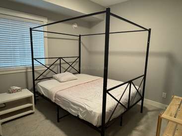 Esta imagen muestra un dormitorio minimalista con una cama con dosel negro, ropa de cama sencilla, una sola almohada, una mesita de noche blanca y paredes y alfombra de color claro.