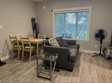 Esta acogedora sala de estar cuenta con un sofá gris, una mesa auxiliar de cristal, un juego de comedor de madera con cojines verdes, una gran ventana, un ventilador de pie y suelos de madera clara.