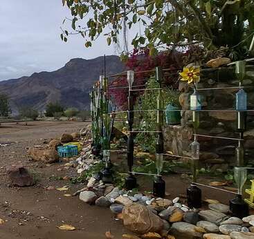 The image shows a creative garden fence made from upside-down glass bottles. Mountains and trees are visible in the background. Decorative stones line the ground.