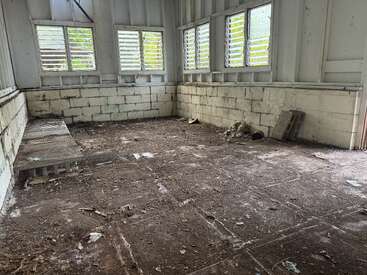This image shows a dirty, abandoned room with concrete block walls, multiple windows with shutters, a wooden bench, debris scattered, and visible deterioration throughout.