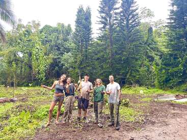 Fünf Menschen stehen lächelnd auf einer üppigen, grünen Waldlichtung, halten Werkzeuge und tragen Arbeitskleidung. Sie sehen glücklich aus, wahrscheinlich nachdem sie ein Gruppenprojekt abgeschlossen haben.