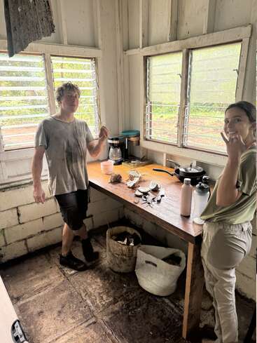 Two people stand in a rustic, sunlit kitchen with open windows, enjoying a snack. Coconut shells, water bottles, and a coffee maker are on the wooden counter.