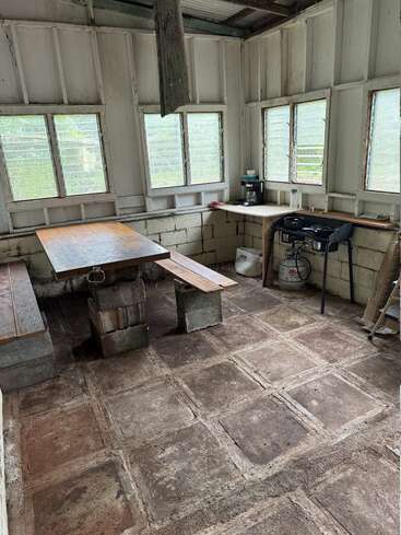 This image shows a rustic, simple kitchen with concrete floors and benches, wooden table, gas stove, coffee maker, and several windows letting in natural light.