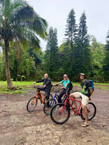 Tres personas en bicicletas de colores se detienen en un camino de tierra rodeado de frondosos árboles tropicales. Parecen felices, disfrutando juntas de una pintoresca aventura al aire libre.