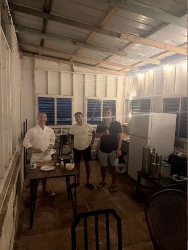 Three men stand in a rustic kitchen with bright lighting. One prepares food, while the others pose and smile. The setting feels casual and relaxed.