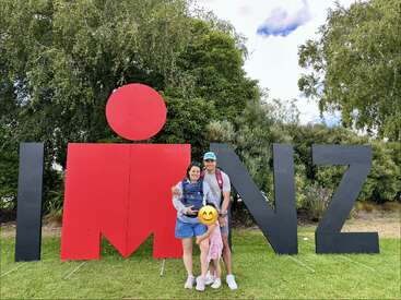 A family stands smiling in front of a large "IM NZ" Ironman sign outdoors, surrounded by lush green trees and grass on a partly cloudy day.
