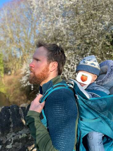 Ein Mann mit rotem Bart trägt ein Baby in einer blauen Rückentrage. Beide sind draußen, warm angezogen, im Hintergrund sind Frühlingsbäume und Sonnenlicht zu sehen.