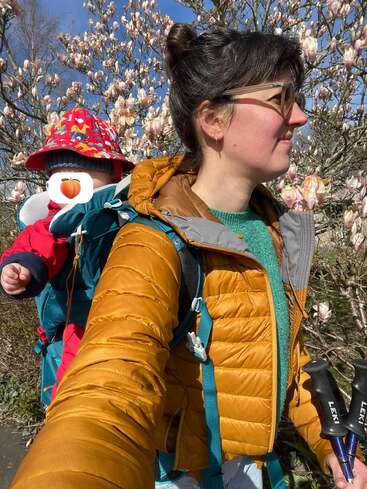 Eine Frau in einer senffarbenen Jacke trägt ein Baby in einer Rückentrage. Beide befinden sich im Freien in der Nähe von blühenden Bäumen und genießen das sonnige Wetter. Das Gesicht des Babys ist verdeckt.