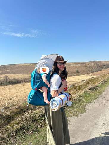 Eine Frau mit Hut und Brille trägt ein Baby in einer blauen Rückentrage, hält eine Decke in der Hand und wandert unter blauem Himmel auf einem sonnigen, ländlichen Weg.