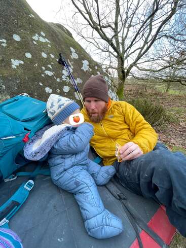 Ein Mann in einer gelben Jacke sitzt auf einer Matte neben einem Felsen und isst gemeinsam mit einem warm eingepackten Kind im Freien unter kahlen Bäumen einen Snack.