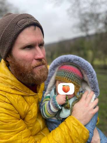 Ein bärtiger Mann in einer gelben Jacke hält ein Baby im Arm, das in bunte Strickkleidung eingepackt ist. Beide tragen Mützen und befinden sich an einem bewölkten Tag im Freien. Das Gesicht des Babys ist von einem Emoji verdeckt.