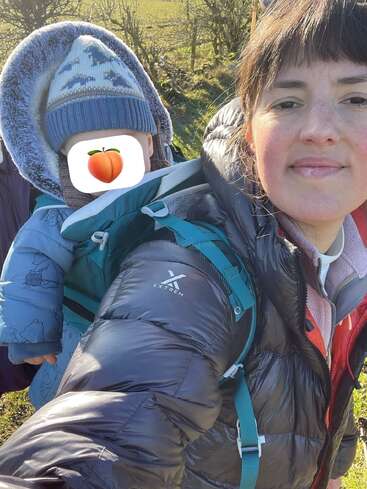 Eine Frau in einer Daunenjacke steht draußen, lächelt und trägt ein warm eingepacktes Kind mit bedecktem Gesicht in einer Babytrage. Es ist ein sonniger, kühler Tag.
