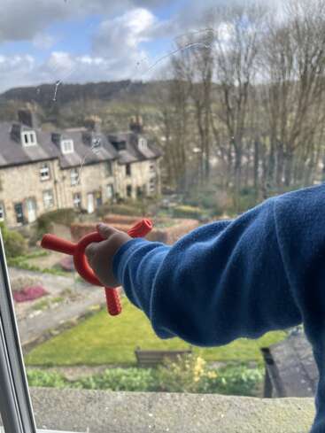 Ein Kind in einem blauen Pullover hält ein rotes Spielzeug an ein Fenster. Draußen sind unter einem teilweise bewölkten Himmel Steinhäuser, ein Garten und Bäume zu sehen.