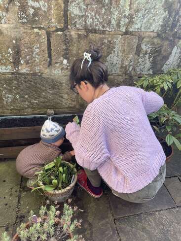 Eine Frau und ein Kind arbeiten gemeinsam im Garten, beide hocken neben Topfpflanzen. Sie sind ganz bei der Sache, umgeben von Steinmauern und viel Grün an einem sonnigen Tag.