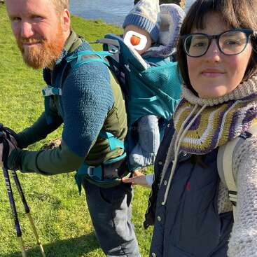 Una escena de una excursión familiar: una mujer se hace un selfi, un hombre con bastones de senderismo lleva a un bebé bien abrigado en una mochila portabebés, con un río y un puente de piedra al fondo.