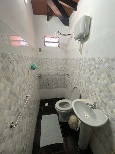 Il s'agit d'une petite salle de bains avec des carreaux muraux à motifs, des toilettes, un lavabo sur pied et une petite fenêtre. Le plafond est orné de poutres apparentes.