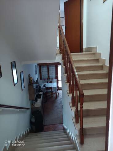 Esta imagen muestra una escalera interior con peldaños de color beige y barandilla de madera, que conduce a un vestíbulo con una puerta abierta. Debajo, se ve una zona de comedor.