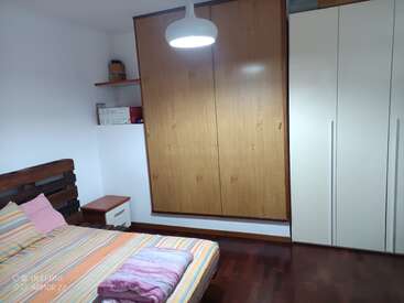 Dormitorio ordenado con colcha a rayas, cabecero de madera, mesilla de noche, amplios armarios, estantería flotante y una lámpara de techo blanca encima. Ambiente acogedor.