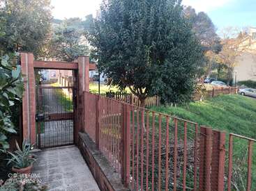 Un camino conduce a una puerta metálica y una valla de ladrillo, con vistas a una zona verde cubierta de hierba con árboles, una maceta y coches aparcados a lo lejos.