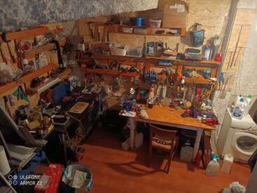 Esta imagen muestra un taller desordenado con estanterías llenas de herramientas, equipos y suministros. También hay un banco de trabajo de madera, una silla y una lavadora.