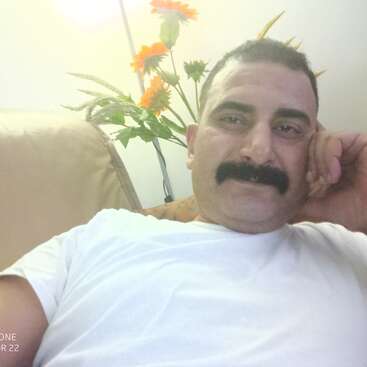 Un hombre con bigote, vestido con camisa blanca, se relaja en un sofá. En el fondo se ven flores brillantes y hojas verdes que crean un ambiente tranquilo.