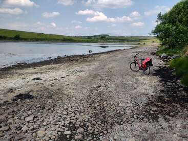 Un vélo rouge avec des sacoches se trouve sur un chemin côtier rocailleux au bord d'une eau calme, entouré d'herbe verte, d'arbres, de collines et d'un ciel partiellement nuageux.