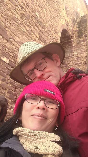 L'image montre un selfie d'un homme et d'une femme devant un mur de briques, la femme portant un bonnet rose et l'homme une veste rouge.