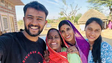 Un alegre grupo de cuatro personas —un hombre y tres mujeres— posa sonriente para hacerse un selfi al aire libre, con casas tradicionales y árboles al fondo bajo un cielo despejado.
