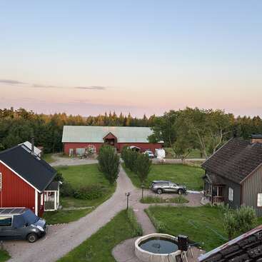 Dieses Bild zeigt eine friedliche Landschaft mit roten Scheunen, grünen Wiesen, Bäumen, geparkten Autos, einem Brunnen und einem Abendhimmel im Hintergrund.