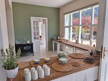 A cozy, sunlit kitchen with large windows, wooden tables set for breakfast, cups, bowls, woven placemats, potted plant, and outdoor view of bright flowers.