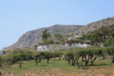 White houses sit among green trees on a hillside, with rugged mountains in the background and a blue sky above, creating a peaceful Mediterranean landscape scene.