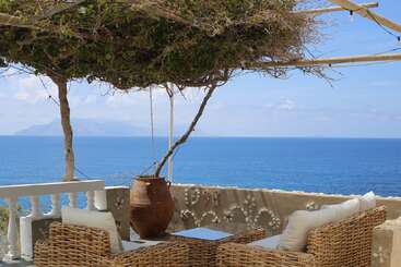 A cozy outdoor seating area with wicker furniture, cushions, a clay pot, and a vine-covered pergola overlooks a beautiful blue sea and distant mountains.