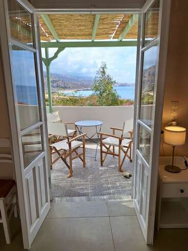 Open French doors reveal a cozy balcony with two chairs and a table, overlooking a scenic seaside landscape with mountains, blue water, and a covered pergola.