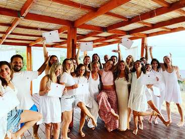 A joyful group of people in white clothing, barefoot, celebrating together with certificates in hand on a wooden deck, possibly after completing a retreat or workshop.