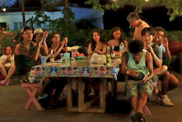 A group of people gathers around a table outdoors at night, some clapping, others relaxing. Drinks, food, and colorful tablecloth create a warm, festive atmosphere.