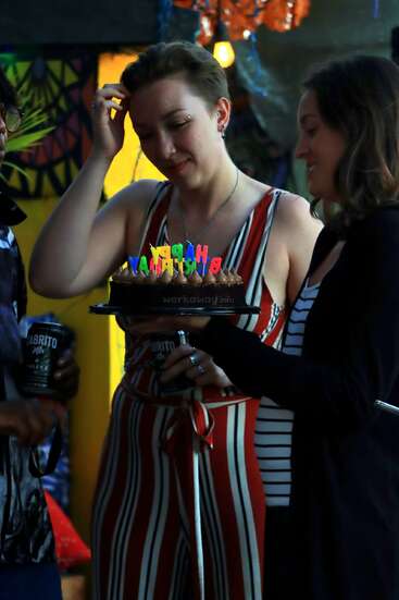 Two women at a birthday party, one holding a chocolate cake with colorful "Happy Birthday" candles, both smiling and enjoying the celebration with friends.