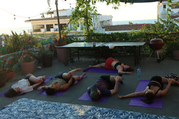 Five people are practicing yoga on purple mats outdoors. Surrounded by plants, they stretch on a shaded terrace with a table, grill, and view of buildings.