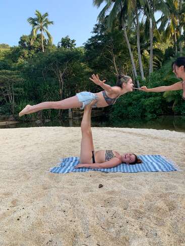 Three women enjoy acro yoga on a sandy beach, surrounded by lush green trees and palm trees. One woman balances another while the third assists playfully.