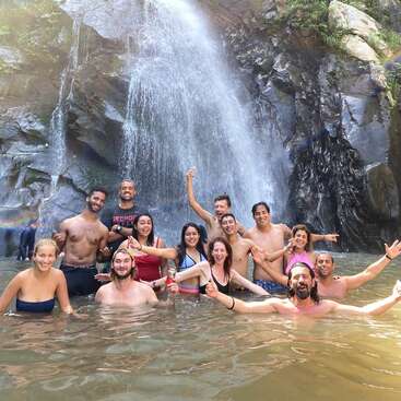 A joyful group of friends poses together in a natural pool beneath a waterfall, smiling and celebrating, surrounded by rocky cliffs and the refreshing spray of water.