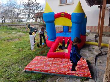 Cinq enfants s'amusent autour d'un château gonflable coloré dans un jardin verdoyant. Certains sautent, glissent et rient, tandis que d'autres les regardent ou s'apprêtent à les rejoindre.