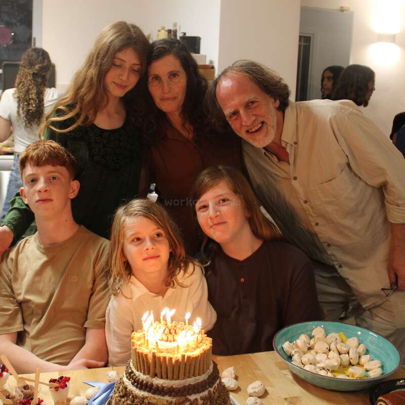 Eine Familie sitzt um einen Tisch herum und feiert einen Geburtstag. Vor ihnen steht eine Torte mit Kerzen, alle lächeln, was eine warme, fröhliche und gemütliche Atmosphäre schafft.
