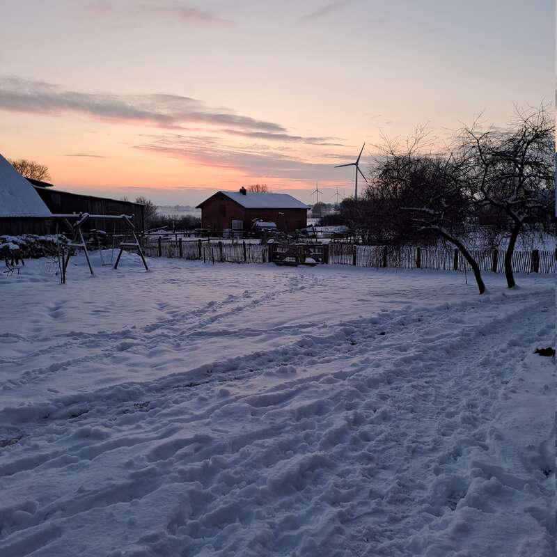 Snow blankets a rural yard at sunset, with a barn, swing set, wind turbines, leafless trees, and a red house creating a peaceful winter landscape.