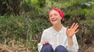 L'image représente une femme aux cheveux longs, vêtue d'une chemise blanche et d'un pantalon noir, debout devant un fond gris, avec une expression neutre et sans objets ou actions visibles.