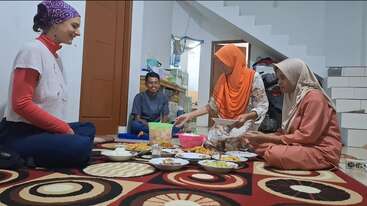 L'image montre un groupe de personnes assises autour d'une table, avec un grand bol de nourriture au centre, entouré d'une nappe et d'autres éléments décoratifs. La scène semble être une réunion sociale ou une célébration.
