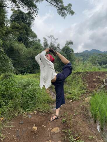 L'image représente une femme debout dans un champ, vêtue d'une chemise blanche, d'un pantalon bleu et de sandales, la jambe droite levée derrière elle et les bras tenant son pied.