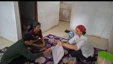 L'image montre un groupe de personnes réunies autour d'une table, avec quelques individus debout à l'arrière-plan, sur fond d'une pièce avec une grande fenêtre.