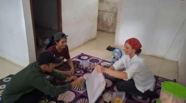The image shows a group of people gathered around a table, with a few individuals standing in the background, set against a backdrop of a room with a large window.